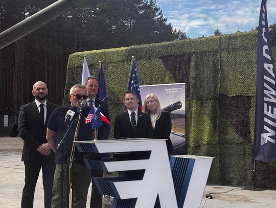 Prezentacja partnerstwa – Niewiadów, Gmina Ujazd Kilka osób stoi za mównicą na zewnętrznej scenie; nad nimi lufa działa, w tle las i banery wydarzenia.