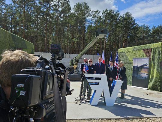 Konferencja prasowa o produkcji amunicji 155 mm Operator kamery filmuje wystąpienie przy mównicy; w tle las, lufa haubicy i flagi państwowe.