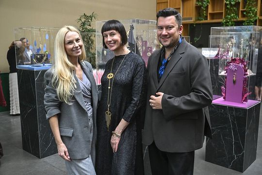 Ekspozycja breloków – spotkanie z gośćmi Trzy osoby pozują na tle gablot z biżuterią ORSKA x PARADYŻ.