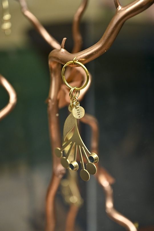 Detal pozłacanego breloka – ORSKA x PARADYŻ Zbliżenie na pozłacany brelok z elementami ceramicznymi zawieszony na metalowej gałęzi.