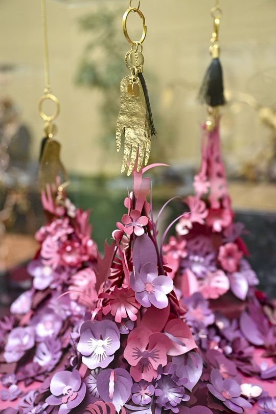 „Dłoń” – symboliczny brelok na kwiatowej instalacji Pozłacana zawieszka w kształcie dłoni nad papierową instalacją z różowo-fioletowych kwiatów.