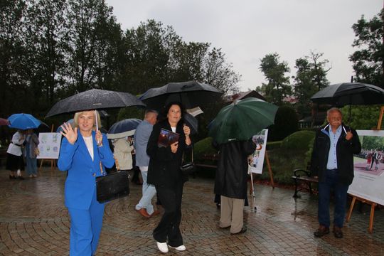 Kobieta macha do aparatu; wokół goście z parasolami i plansze wystawy w parku.