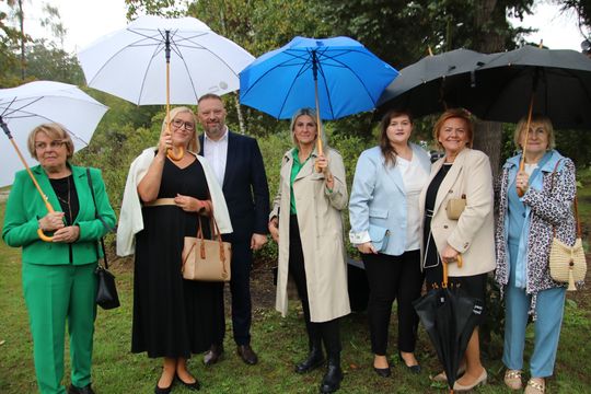 Siedem osób w eleganckich strojach stoi na trawie pod parasolami, pozując do zdjęcia podczas deszczowej uroczystości plenerowej.