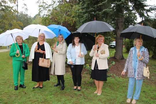 Siedem osób ustawionych w rzędzie z parasolami na tle drzew; pamiątkowe zdjęcie wykonane w deszczu.