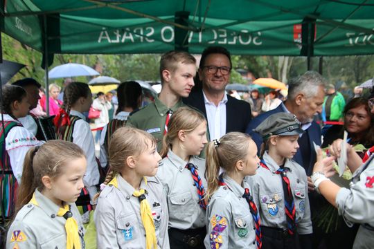 Dzieci w mundurach harcerskich z chustami stoją pod namiotem obok dorosłych gości podczas plenerowej uroczystości.