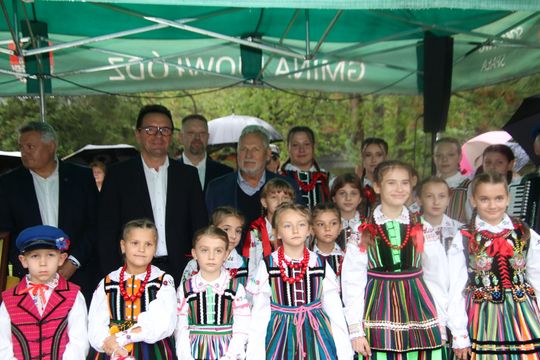 Grupa dzieci w kolorowych strojach ludowych stoi frontem do aparatu, za nimi dorośli pod namiotem.