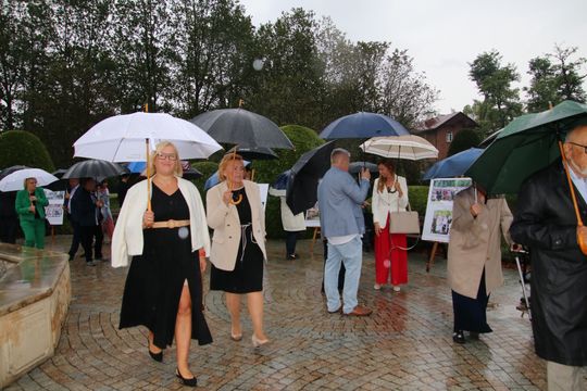 Elegancja w kroplach deszczu Uczestnicy z parasolami idą alejką parku, oglądając plenerową ekspozycję o dożynkach.