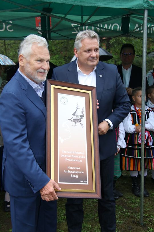 Prezentacja wyróżnienia – kadr z bliska Dwie osoby pokazują do kamery oprawiony dyplom z napisem „Honorowi Ambasadorowie Spały”; w tle uczestnicy w strojach ludowych.