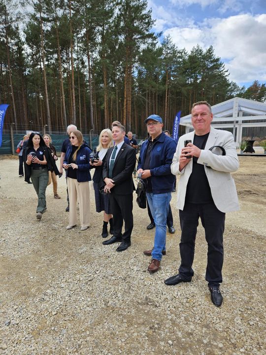 Grupa osób stoi na żwirowym placu przy lesie podczas wydarzenia branżowego; w tle namiot i banery.