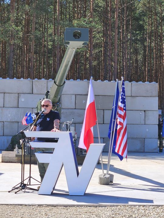 Mówca przy mównicy z logo, obok flagi Polski i USA; za nim widać lufę haubicy i ścianę ochronną.