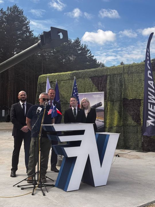 Kilka osób stoi za mównicą na zewnętrznej scenie; nad nimi lufa działa, w tle las i banery wydarzenia.