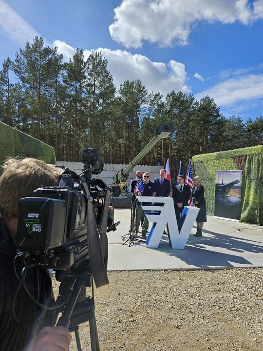 Operator kamery filmuje wystąpienie przy mównicy; w tle las, lufa haubicy i flagi państwowe.