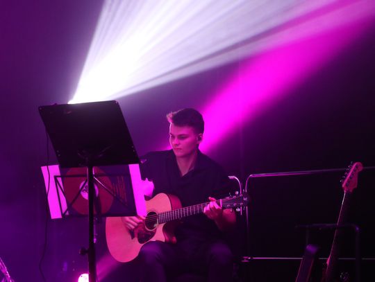 Gitarzysta akustyczny przy pulpicie nut, intensywne światło w kolorze magenta.