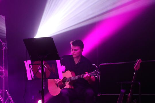 Młody gitarzysta na scenie PCAS Gitarzysta akustyczny przy pulpicie nut, intensywne światło w kolorze magenta.