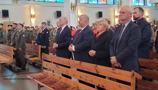 Przedstawiciele władz samorządowych i służb mundurowych stoją w ławkach podczas mszy rocznicowej.